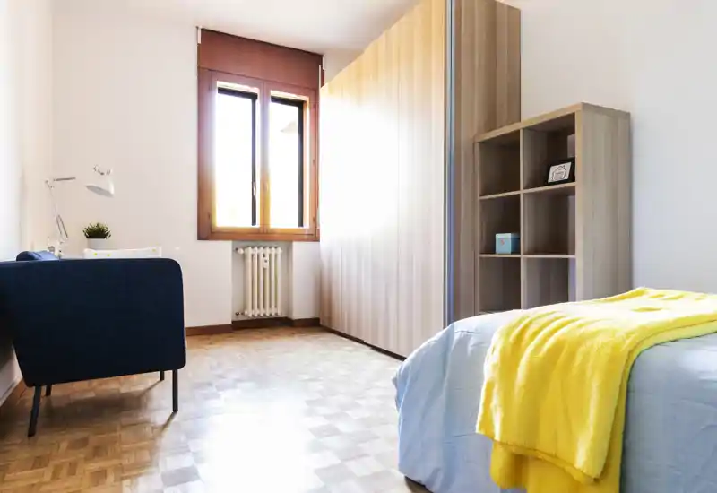 Bright, tidy bedroom with a light-wood wardrobe and open shelving. The single bed in the foreground is dressed in soft linens and a cheerful yellow throw, and a window provides warm natural light.
