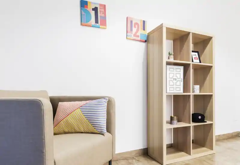 Bright, minimal living area corner with a beige sofa, colorful striped cushion and a tall wooden cubed shelving unit with decorative items — a tidy, modern spot for relaxing.