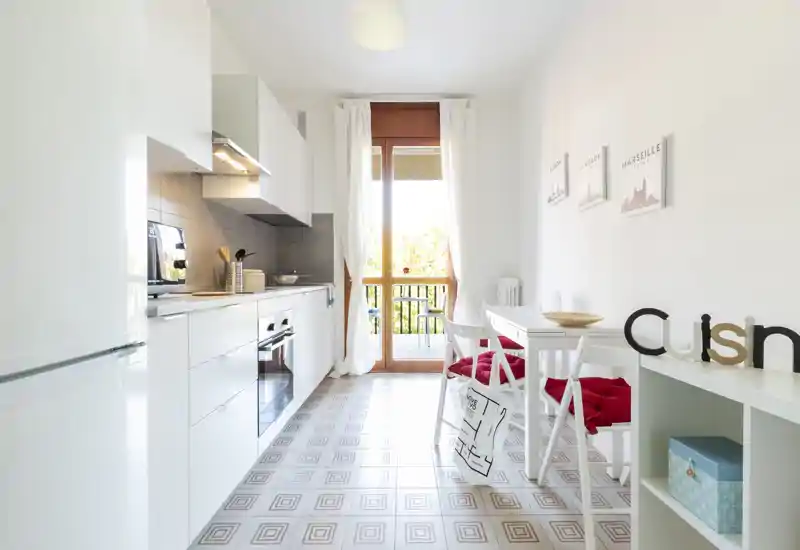 Bright modern kitchen with integrated appliances, white cabinets and a small dining area by a glass door leading to a balcony — clean, well-lit space ideal for short-term rentals.