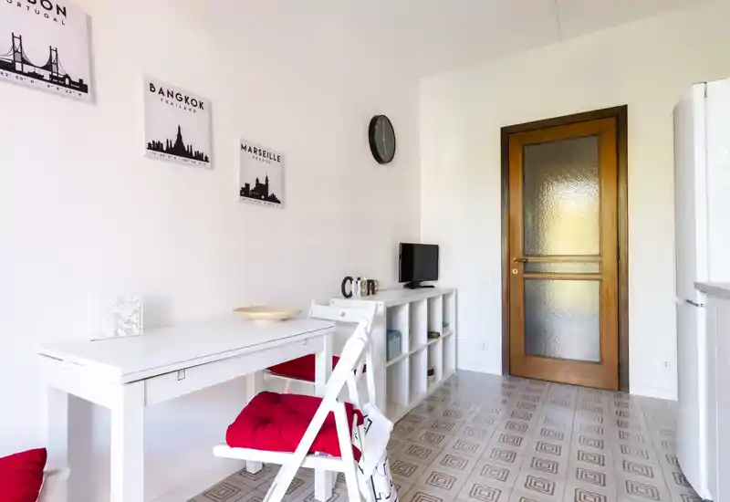 Bright, compact dining area with a white table and red-cushioned folding chairs, decorative wall prints and a low shelving unit — ideal for a small apartment or studio.
