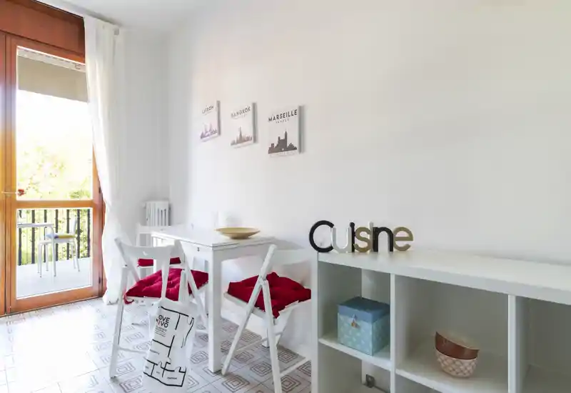 Bright, compact dining corner with a small white table, two chairs with red cushions and shelving; natural light from a balcony door creates an airy feel.