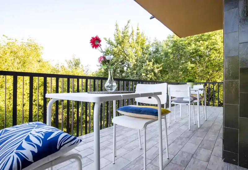 Sunny furnished balcony with a small dining table, white chairs and colorful cushions overlooking green trees — ideal for morning coffee or evening relaxation.