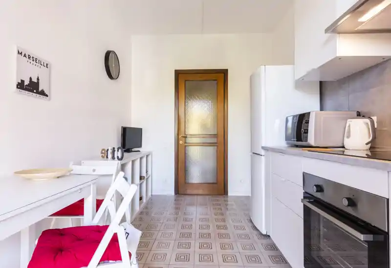 Compact, modern kitchenette with white cabinets, full-size fridge, oven and a small dining area with red-cushioned chairs; bright and neatly styled for short-term stays.