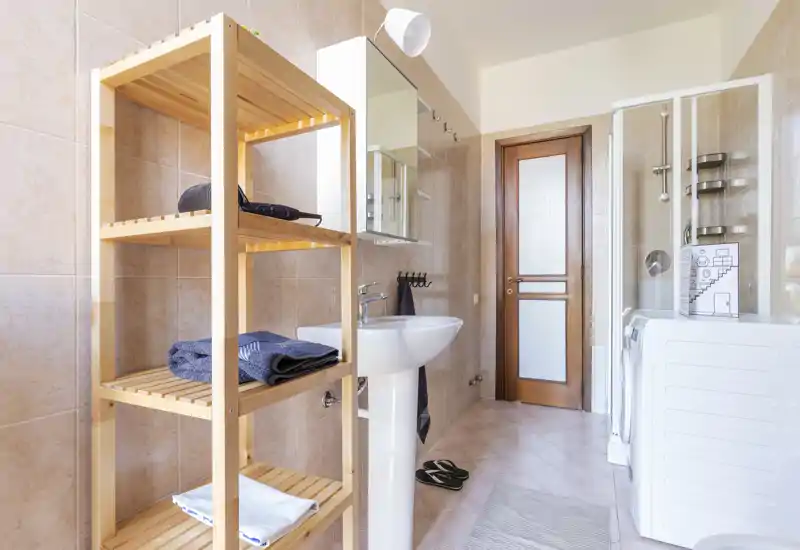 Bright, modern bathroom with a pedestal sink, glass shower enclosure and a washing machine; wooden shelving provides storage and towels.