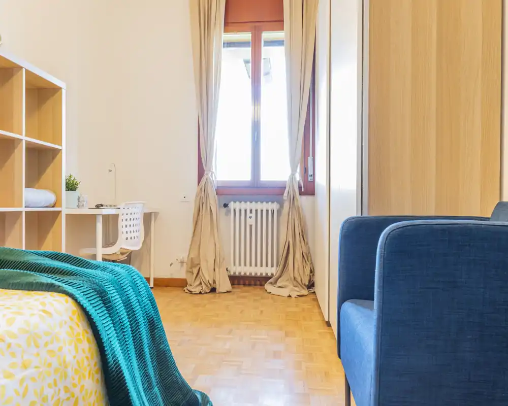 Bright, well-styled bedroom featuring a patterned duvet and teal throw, with a desk by the window and warm wooden flooring.
