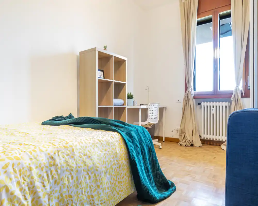 Bright, well-staged bedroom with a patterned bedspread and teal throw, a small desk and shelving by the window — comfortable and naturally lit.