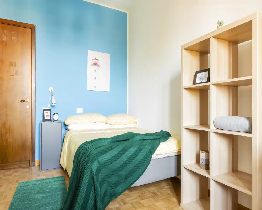 Bright, neatly staged bedroom with a double bed against a turquoise accent wall, cozy textiles and a light wooden shelving unit providing storage.