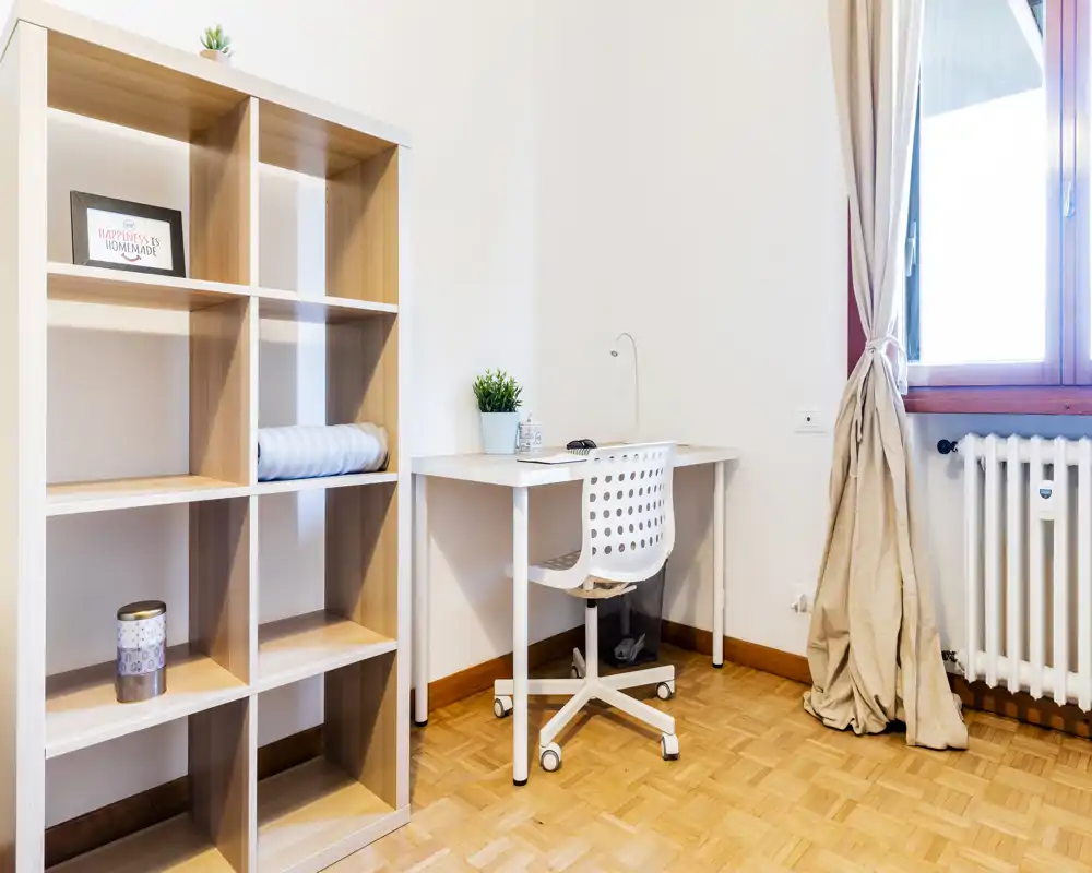 Small, bright room corner with a white study desk, ergonomic chair and open shelving — ideal for a student or home office nook with natural window light.