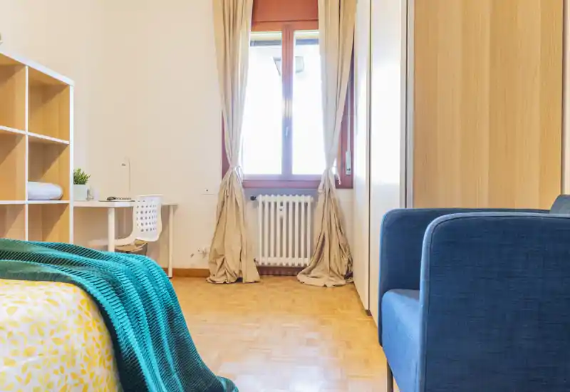 Bright, well-styled bedroom featuring a patterned duvet and teal throw, with a desk by the window and warm wooden flooring.