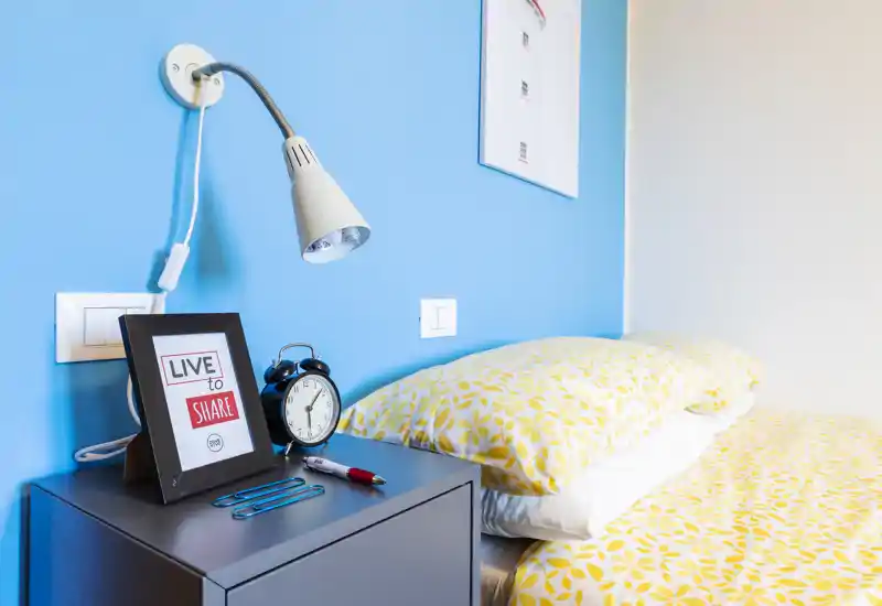 Bright, modern bedroom corner with a patterned yellow duvet, bedside table, alarm clock and a wall-mounted reading lamp against a vivid blue accent wall.