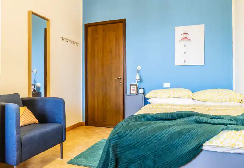 Bright, stylish bedroom with a comfortable double bed against a teal accent wall, layered bedding and a cozy armchair — ideal for a relaxed stay.