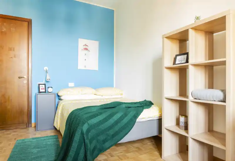 Bright, neatly staged bedroom with a double bed against a turquoise accent wall, cozy textiles and a light wooden shelving unit providing storage.