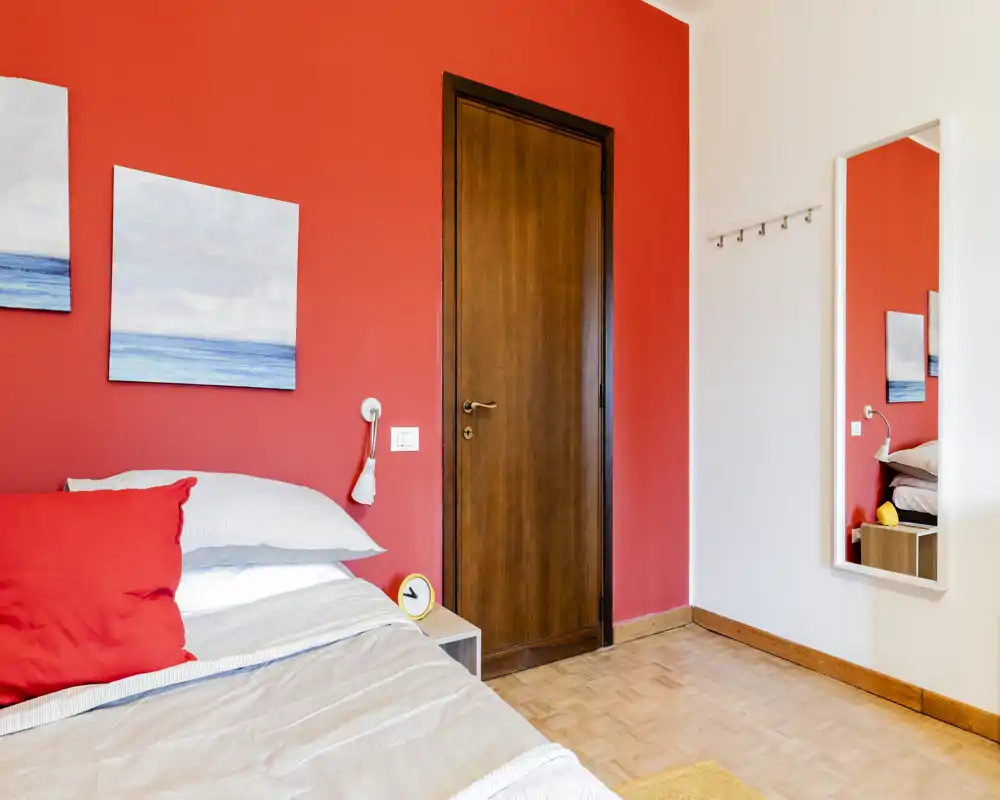 Bright, modern bedroom with a bold red accent wall, neatly made bed with layered pillows and a small bedside table featuring a round yellow clock. A full-length mirror and minimalist coastal artwork complete the clean, contemporary look.