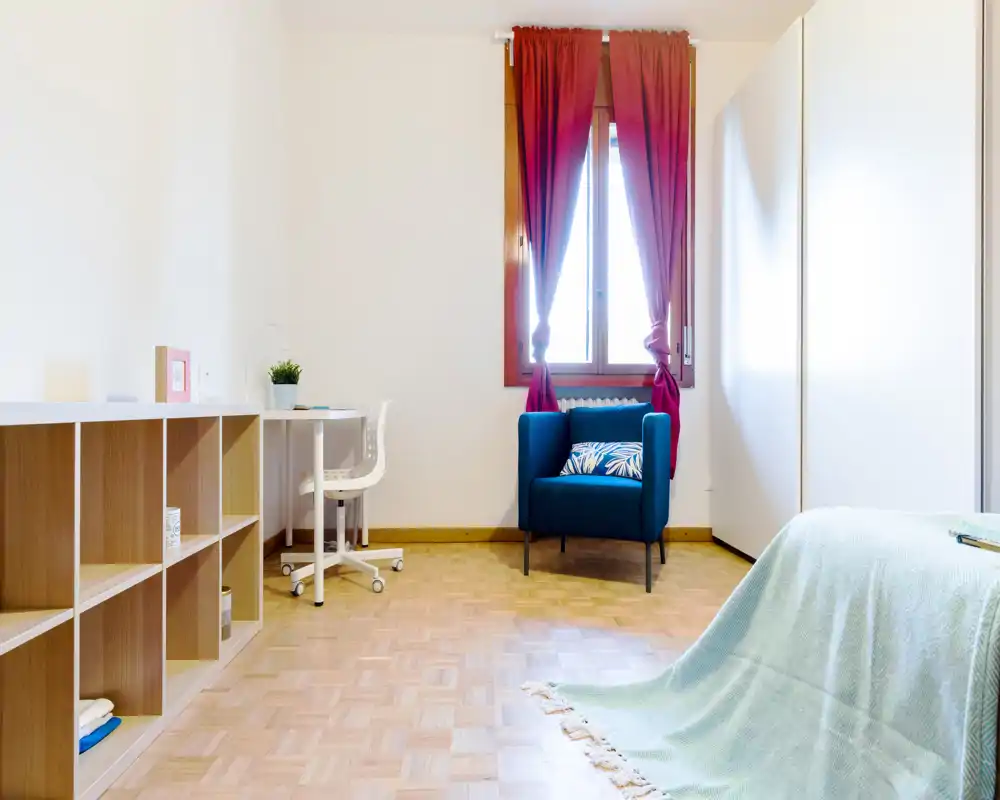 Bright, simply furnished bedroom with a single bed draped in a light throw, a cozy blue armchair by the window and a small desk with shelving — ideal for a student or single occupant.