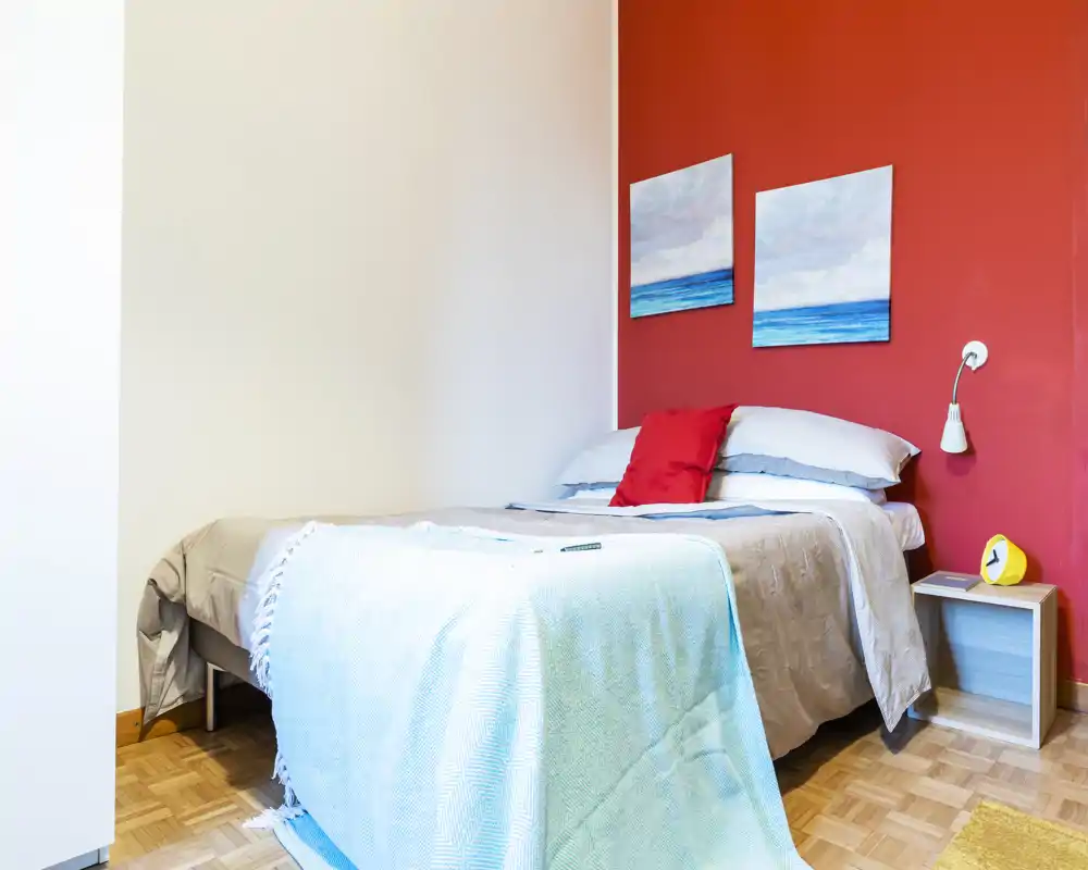 Bright, neatly made bedroom with a double bed against a red accent wall, decorative seascape paintings, a nightstand and soft textiles creating a welcoming atmosphere.