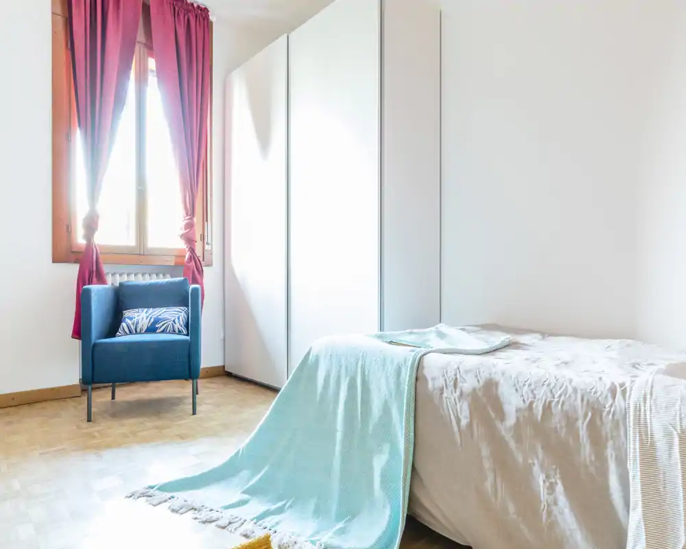 Bright, simply styled bedroom with a neatly made single bed, turquoise throw and a blue accent armchair by the window. Large white wardrobe and natural light create an airy, uncluttered feeling ideal for listing photos.
