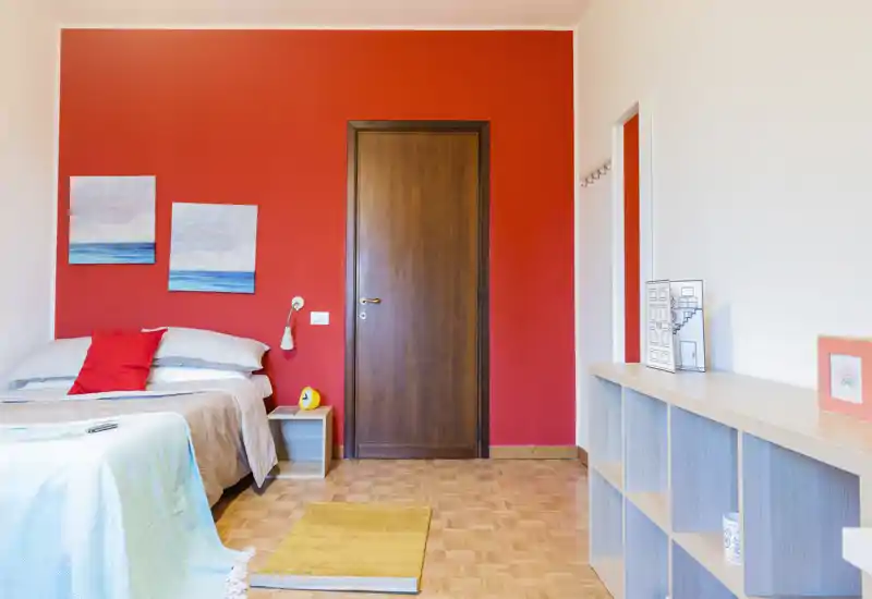 Bright, stylish bedroom with a double bed against a warm red accent wall, bedside table, and shelving unit — ideal for a cozy rental stay.