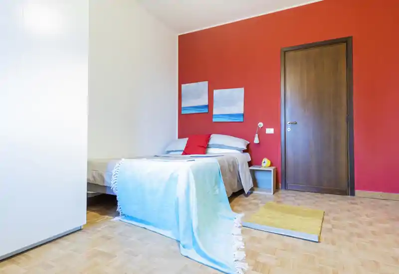 Bright, modern bedroom with a double bed dressed in light blue linens, decorative pillows and a contrasting red accent wall. The room features a wooden door, small bedside shelf and parquet flooring for a cozy, well-styled feel.