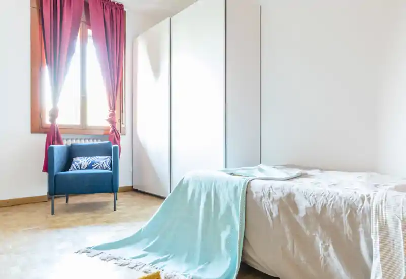 Bright, simply styled bedroom with a neatly made single bed, turquoise throw and a blue accent armchair by the window. Large white wardrobe and natural light create an airy, uncluttered feeling ideal for listing photos.