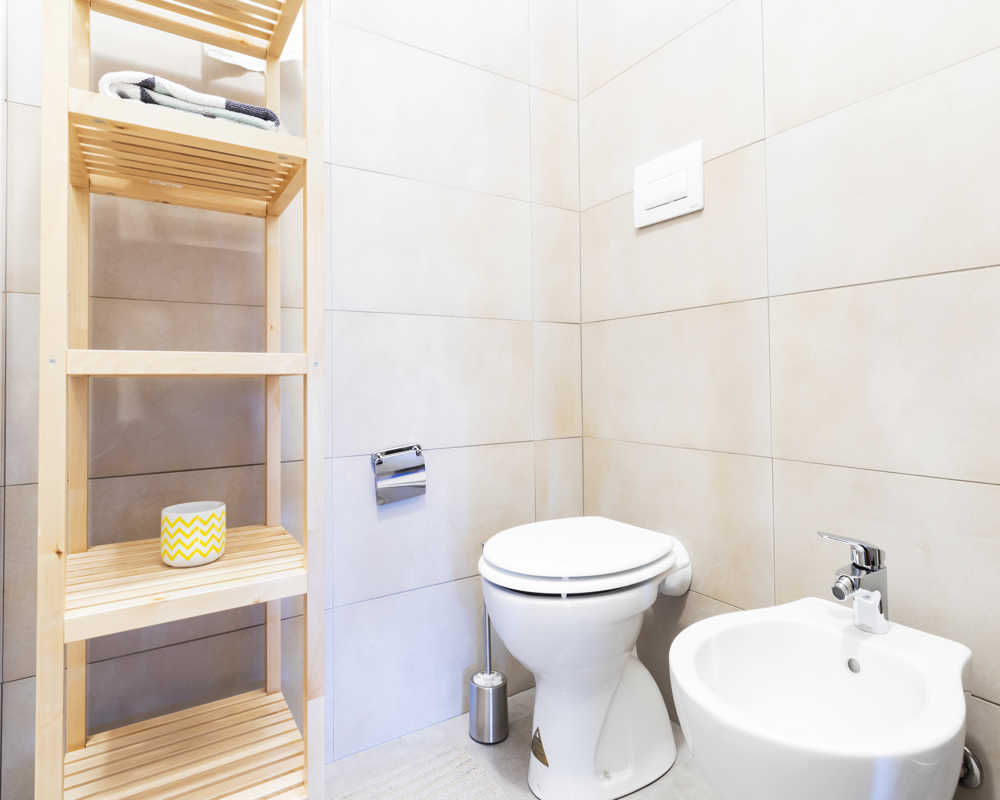 Compact modern bathroom with white toilet, bidet and a wooden shelving unit; bright tiles and contemporary fittings create a clean, fresh feel.