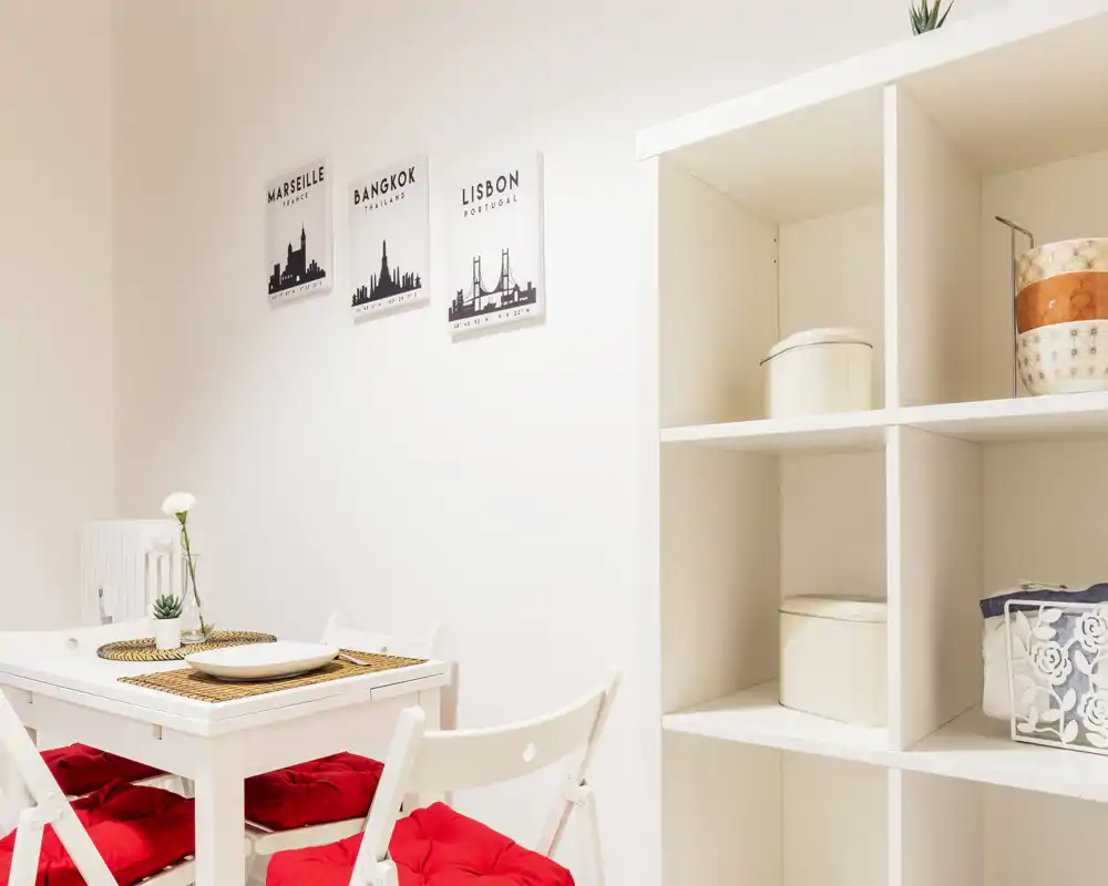 Bright, modern dining corner with a small white table, red seat cushions and minimalist shelving — ideal for cozy meals or a compact apartment dining area.