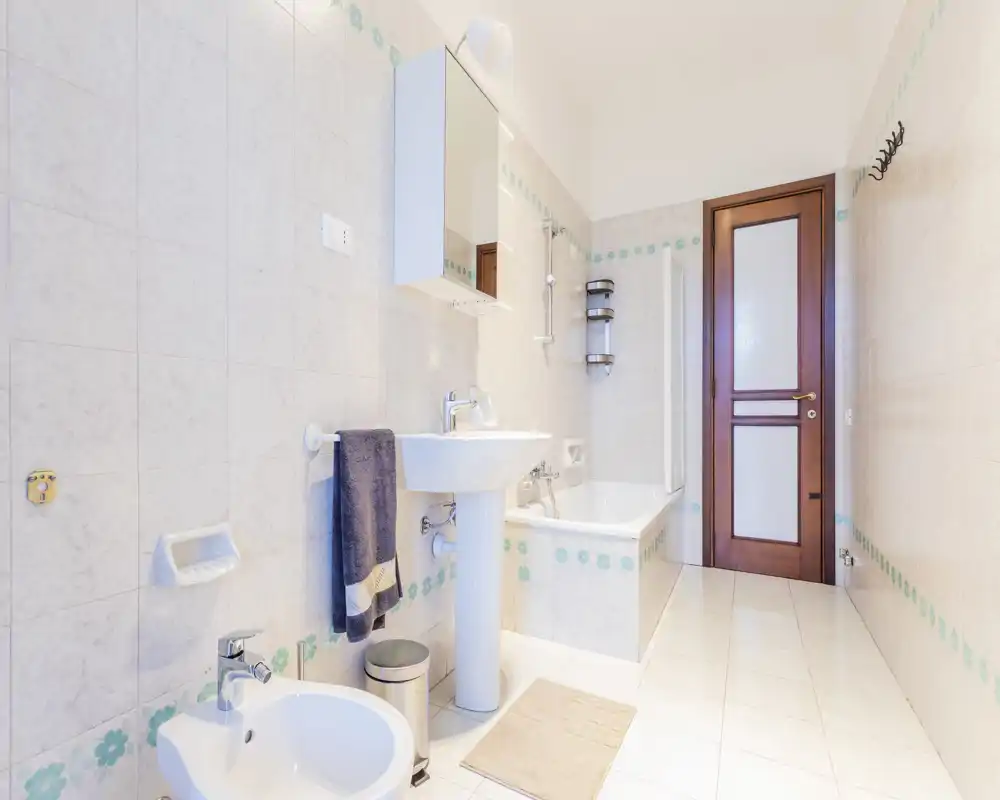 Bright, well-kept bathroom with bathtub, pedestal sink, bidet and mirrored cabinet — neutral tiles and soft accents create a clean, airy feel.