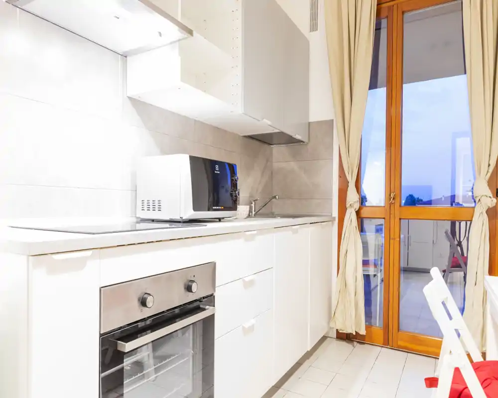 Bright modern kitchen with white cabinetry, built-in oven, microwave and a glass door leading to a balcony—clean, minimalist and well lit.