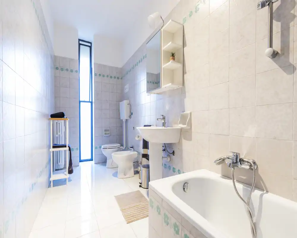 Bright, well-lit bathroom with a bathtub in the foreground, a sink with mirror cabinet, toilet and bidet visible; tiles with subtle decorative border and a tall narrow window providing natural light.