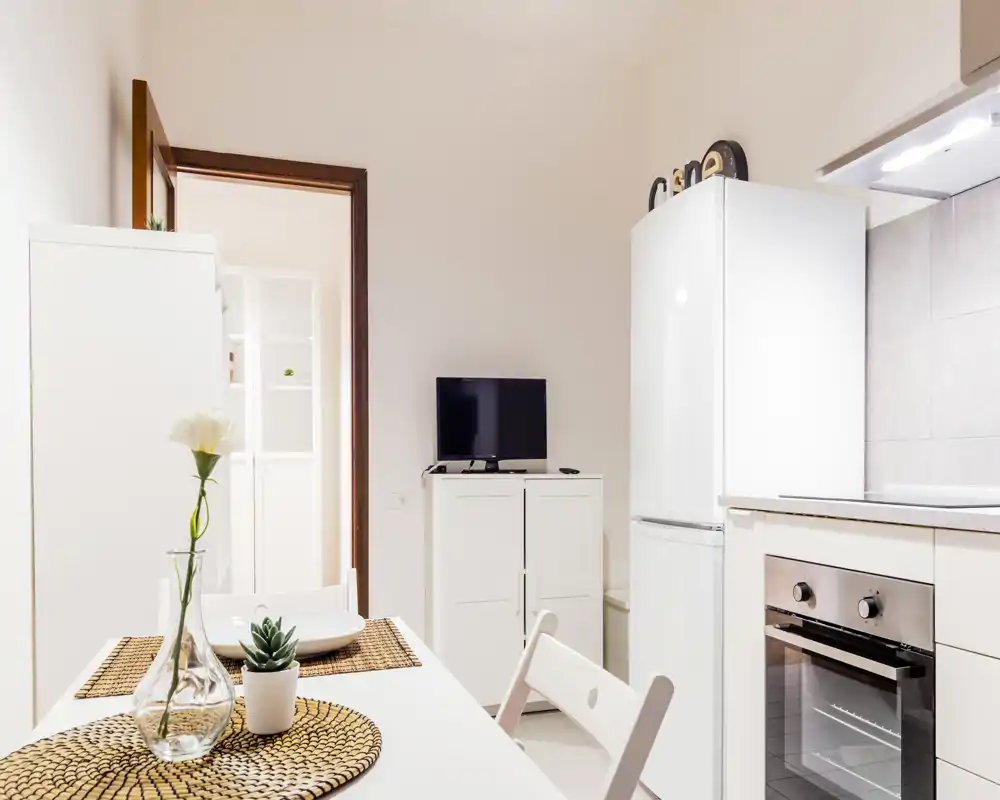 Bright modern kitchenette with a small dining table set for two, decorative placemats, a vase and compact appliances including a fridge and oven.
