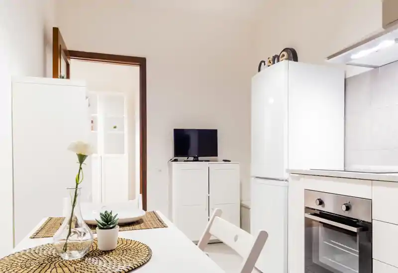 Bright modern kitchenette with a small dining table set for two, decorative placemats, a vase and compact appliances including a fridge and oven.
