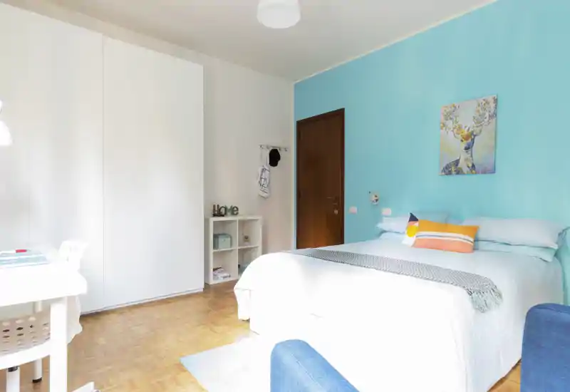 Camera da letto luminosa e ben arredata con letto matrimoniale con biancheria bianca, cuscini colorati, parete accent turchese e ampio armadio — perfetta per annunci.
