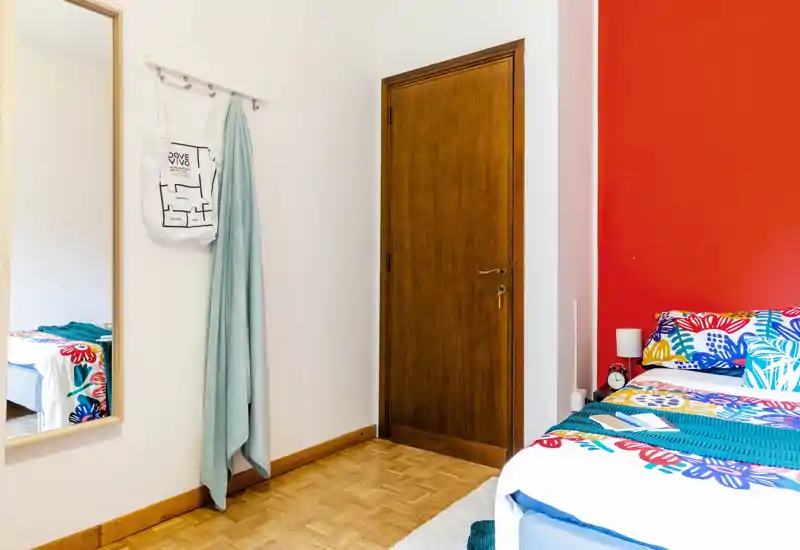 Bright, tidy bedroom corner with colorful bedding and a wooden door; a mirror and hanging textiles add depth and personality.