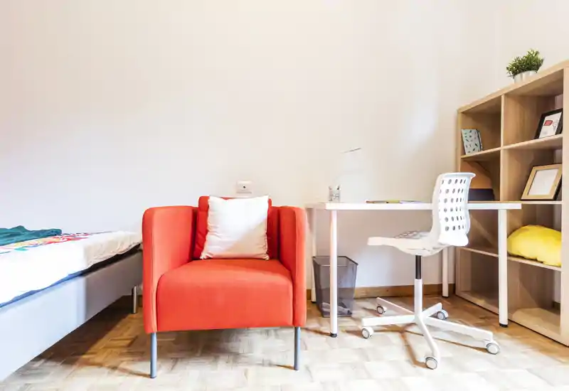 Bright, modern bedroom corner featuring a single bed at left, a vibrant orange armchair, and a compact desk with shelving — ideal for a student or short-term rental.