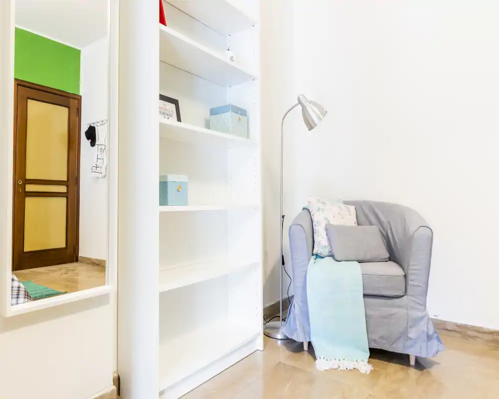 Bright corner with a single upholstered armchair, floor lamp and shelving — a compact living/reading nook with soft textiles and neutral decor.