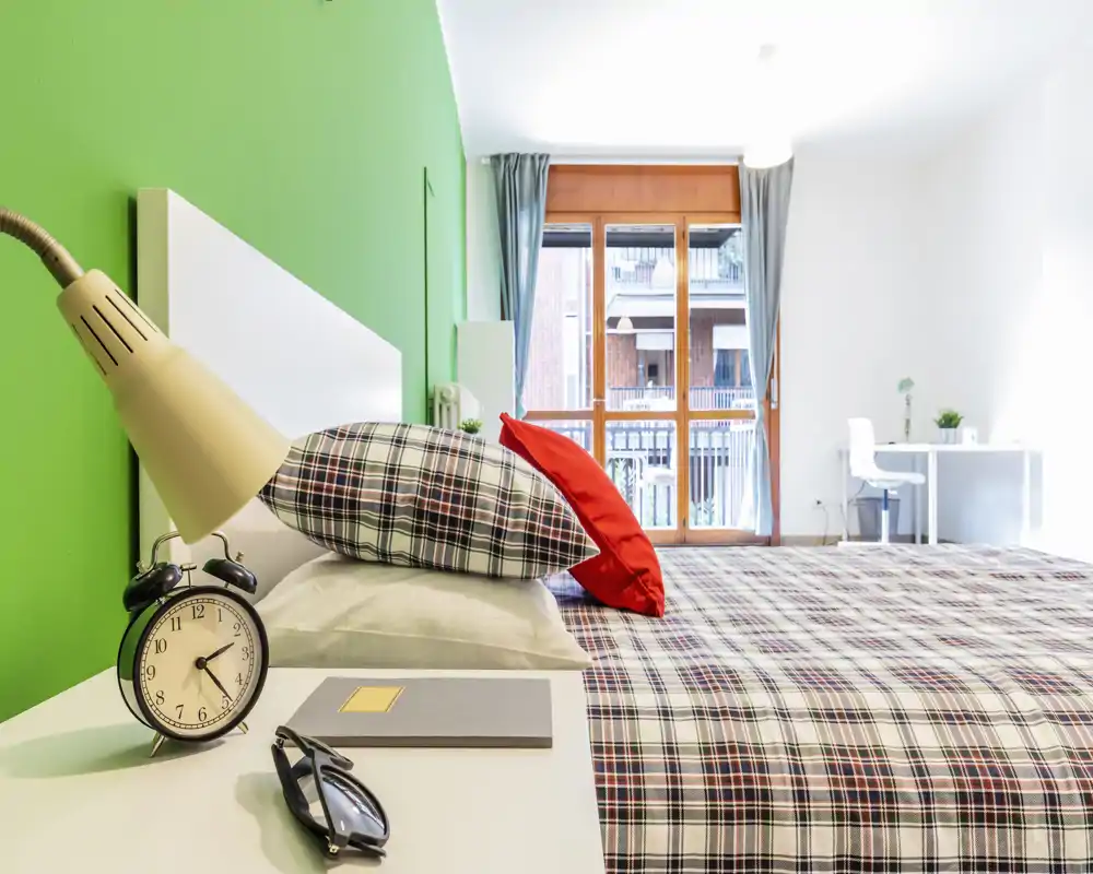 Bright, modern bedroom with a large bed dressed in patterned linens, colorful cushions and a bedside table with lamp and alarm clock. Large window/door brings abundant natural light and a small desk is visible near the window.