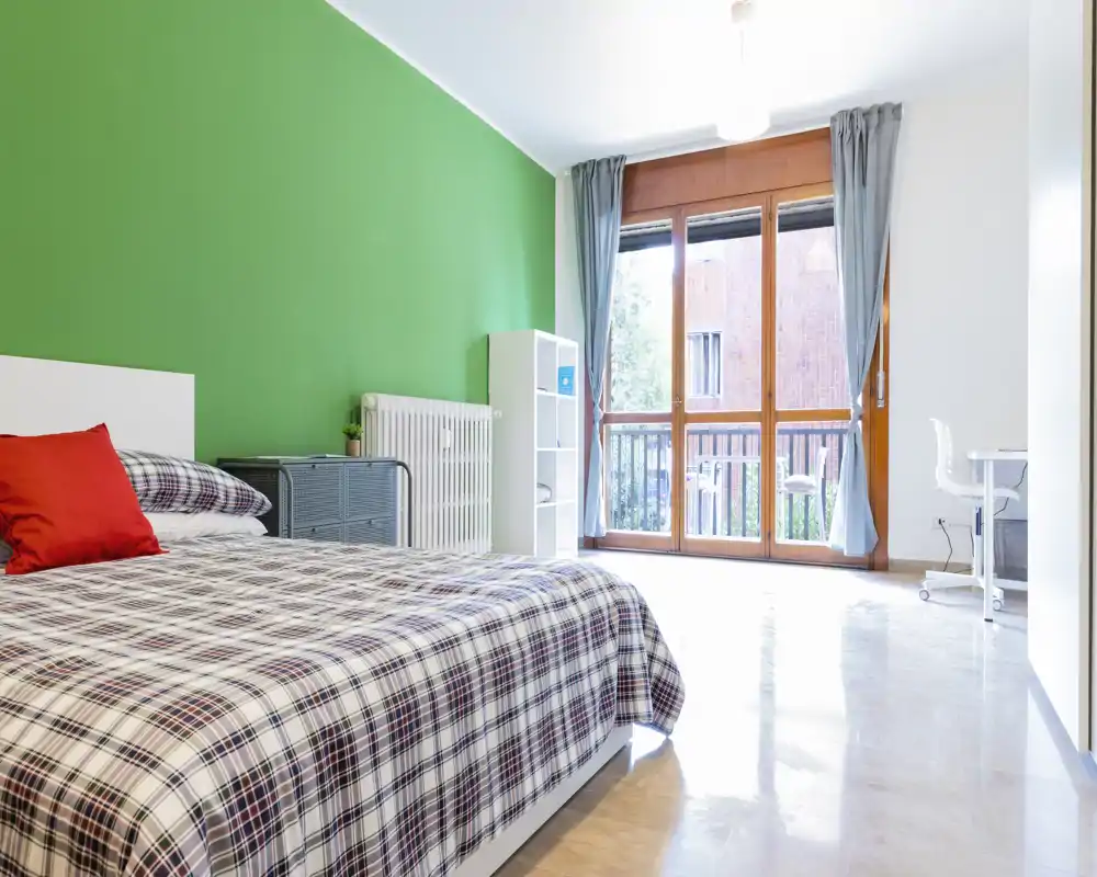 Bright, well-staged bedroom with a double bed covered in checked bedding, large window/door letting in abundant natural light and a small desk by the window.