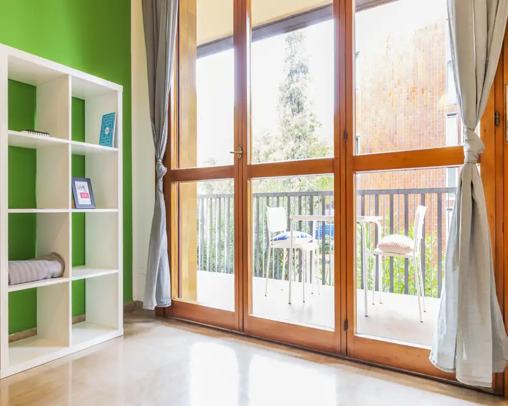 Bright room with large wooden-framed glass doors opening onto a small balcony furnished with chairs — plenty of natural light and a leafy view.