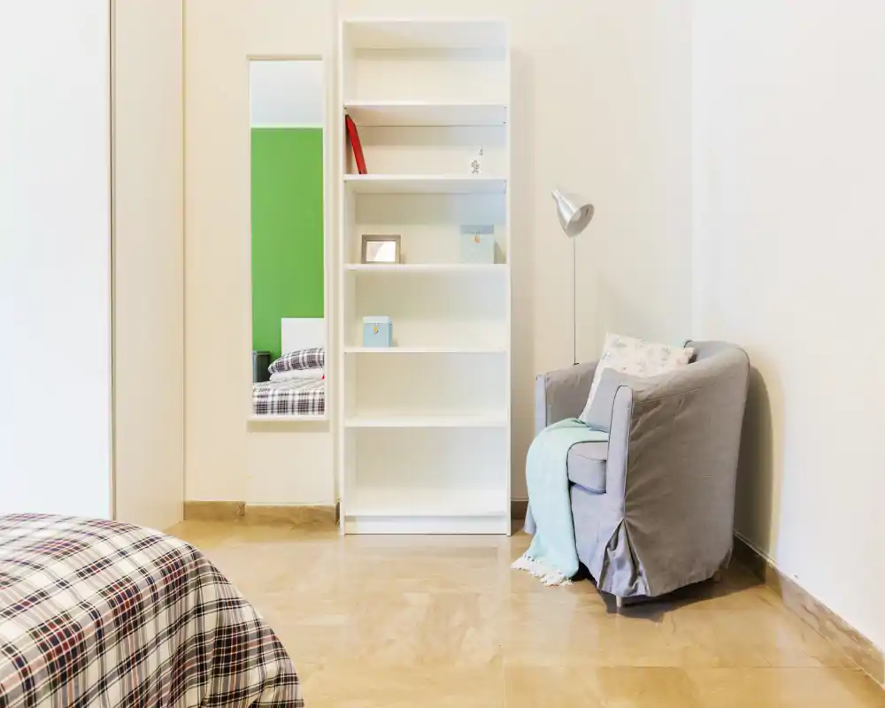 Bright, tidy bedroom corner with a patterned bed visible in the foreground, a white shelving unit and a comfortable armchair with a throw — simple, modern styling ideal for a rental listing.