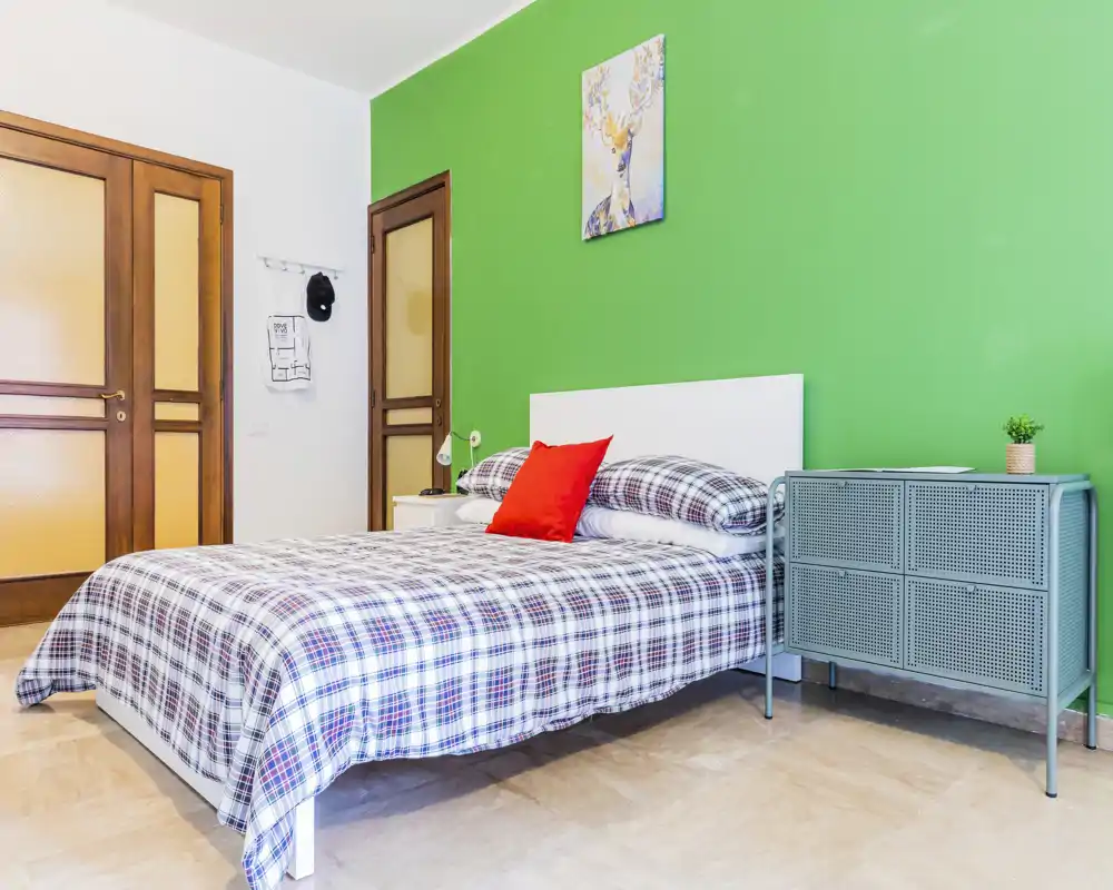 Bright, modern bedroom with a double bed dressed in checked linens, a bold green accent wall and simple bedside storage — well lit and comfortably styled for short-term rental photos.