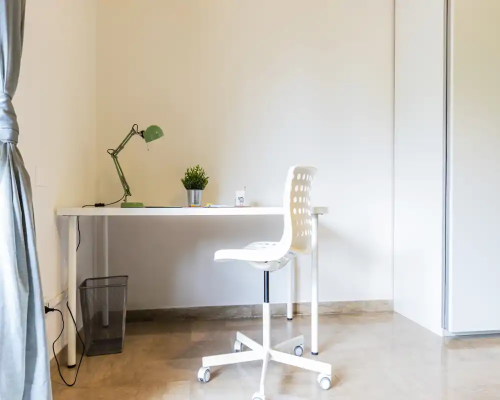 Bright, minimalist workspace with a white desk, adjustable green lamp, small plant and a white swivel chair on wheels. The scene suggests a small home office or study corner in a neutral, uncluttered room.