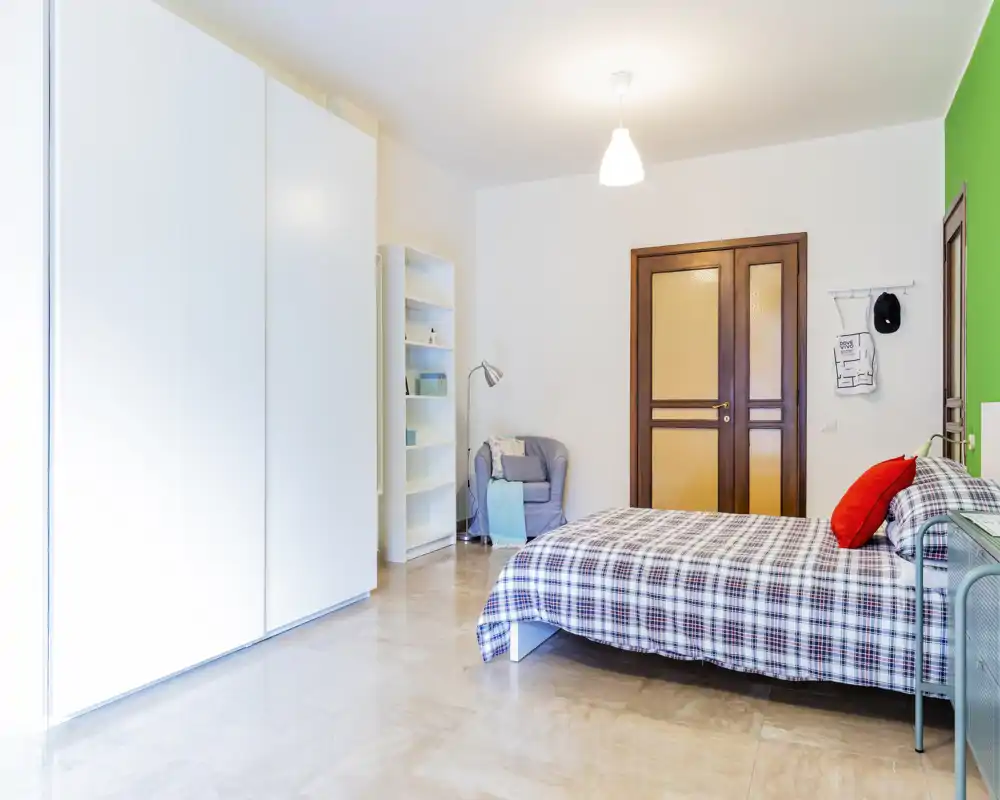 Bright, modern bedroom with a double bed in plaid bedding, a large white wardrobe and a cozy armchair — well-lit and tastefully furnished.