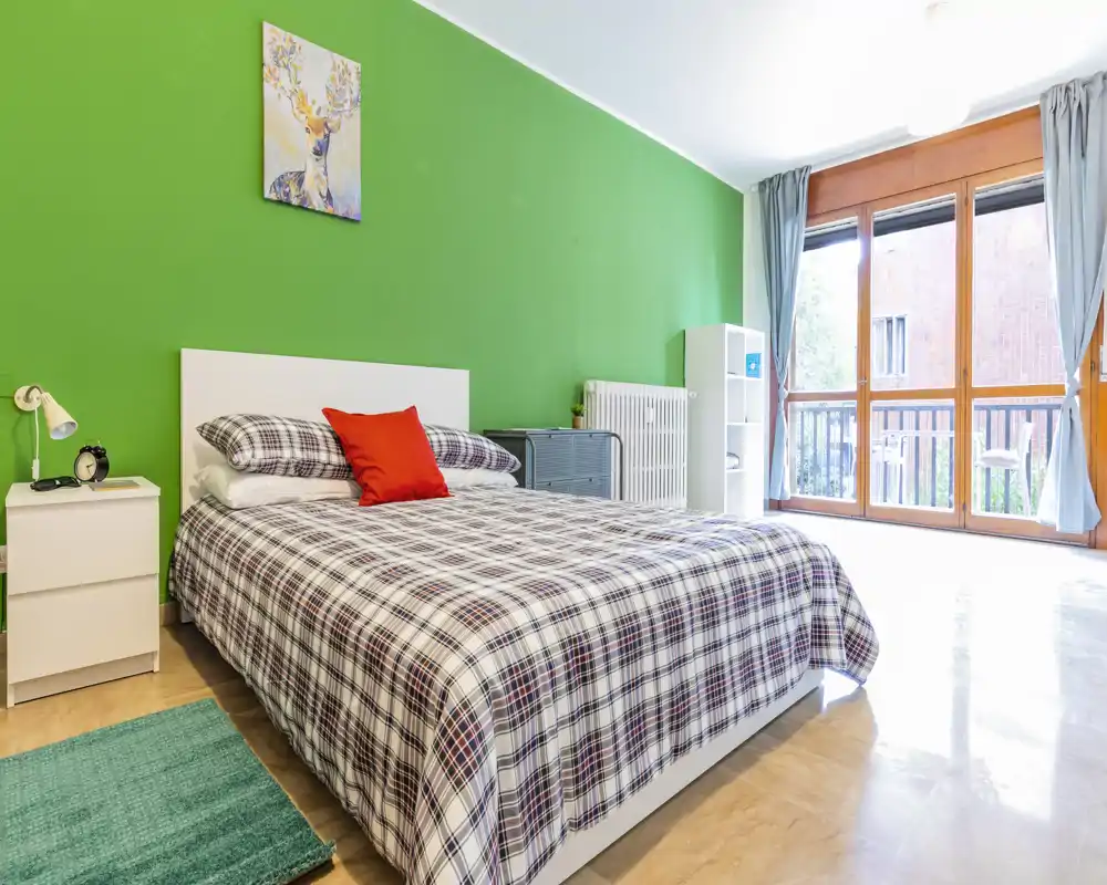 Bright, well-styled bedroom with a double bed against a vibrant green accent wall and large windows providing abundant natural light.