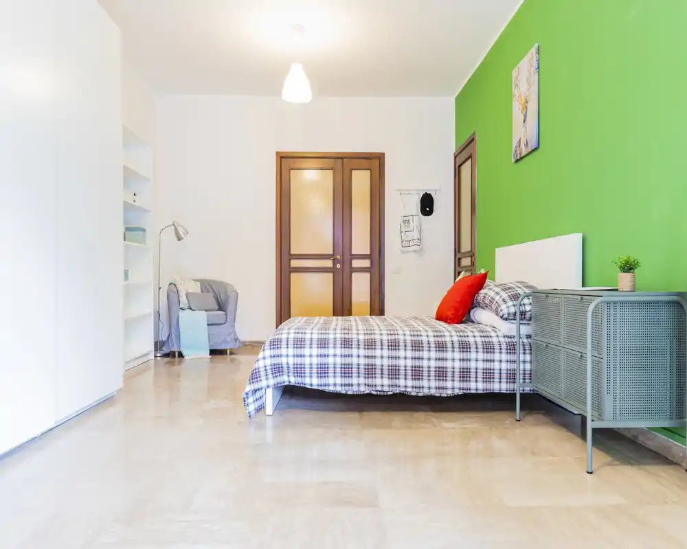 Bright, well-styled bedroom with a double bed dressed in checked linens, a green accent wall and a small bedside console. The room feels airy and modern, ideal for a rental listing photo.