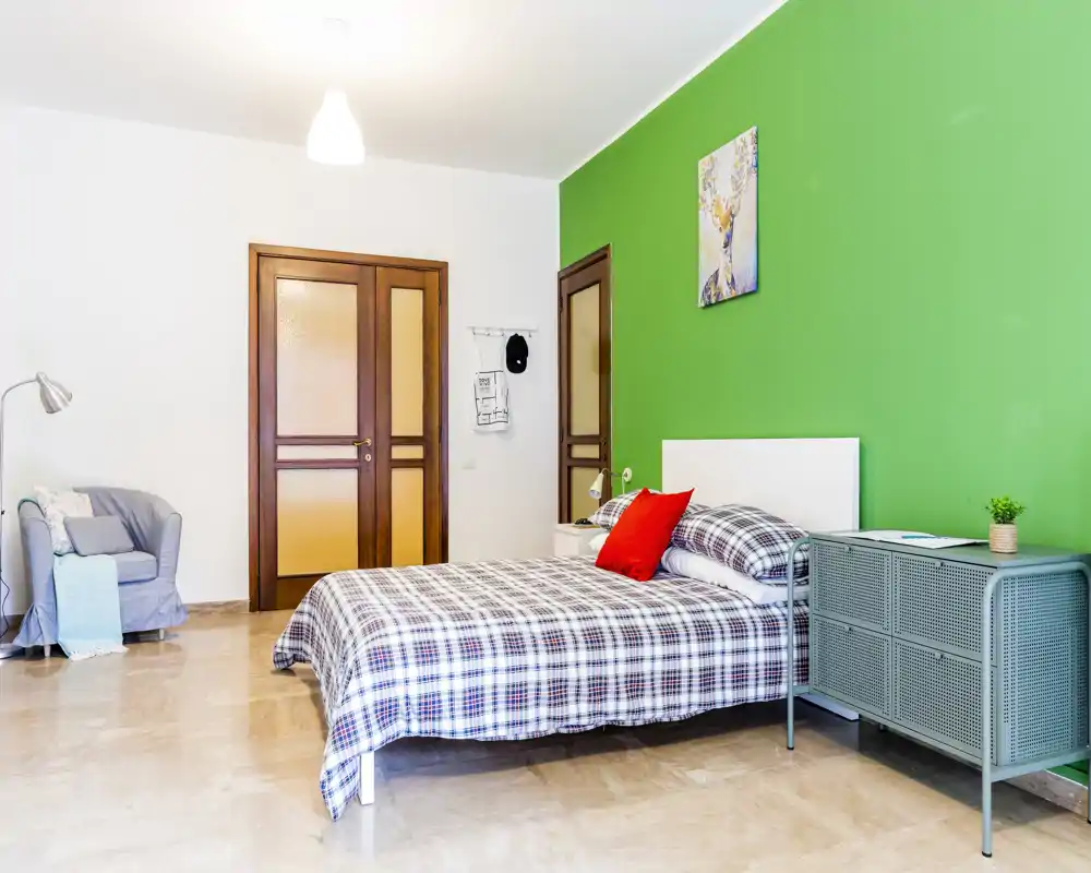 Bright, modern bedroom with a double bed dressed in checked linens, a green accent wall and a cozy armchair — ideal for rental photos highlighting comfort and color.
