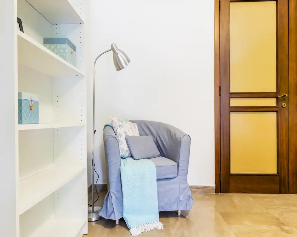 A bright, cozy corner living area featuring a comfortable armchair with cushions and a throw, a standing lamp, and shelving — ideal for reading or relaxing.
