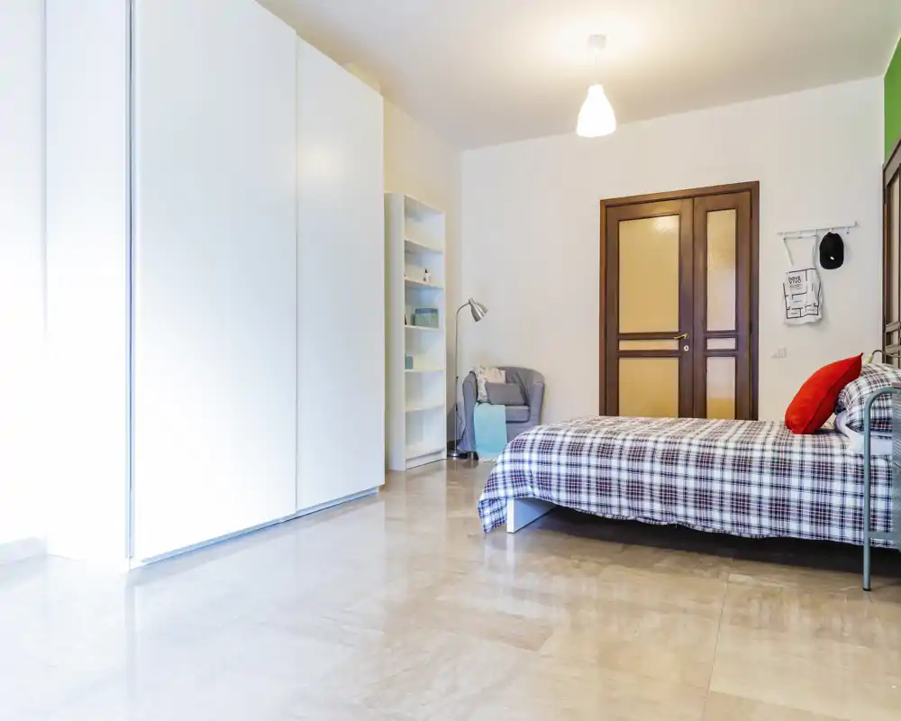 Bright, modern bedroom with a single bed dressed in checked bedding, a large white wardrobe, shelving and a cozy armchair near the door — clean, minimal and airy.