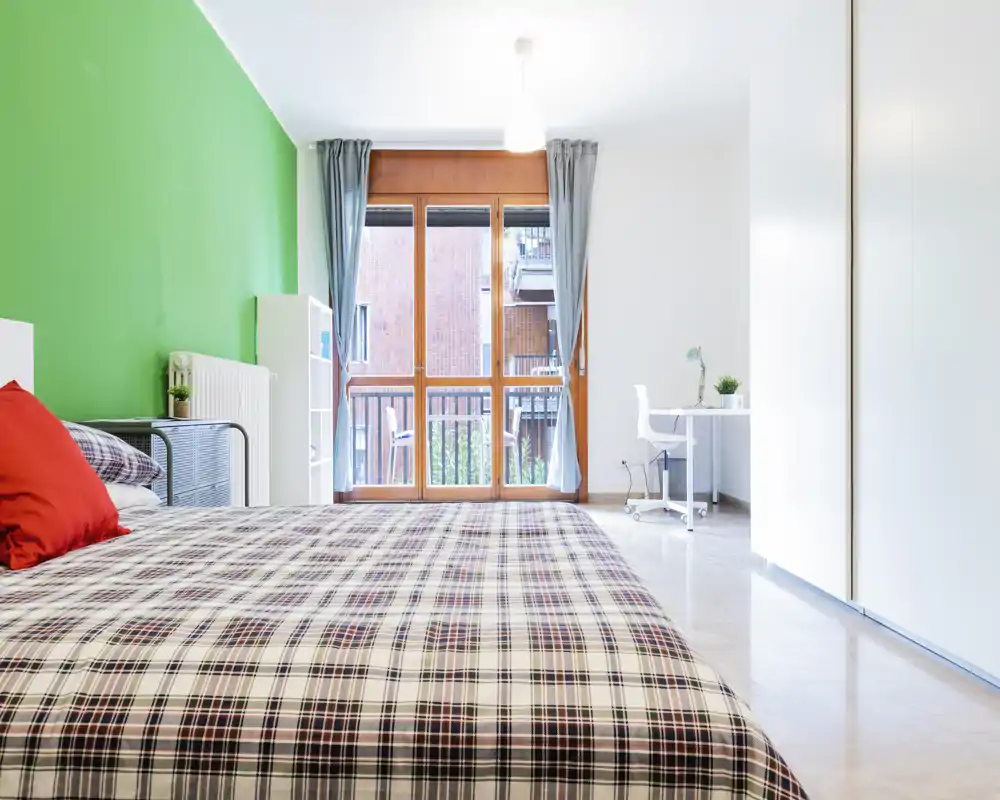 Bright, modern bedroom with a large bed in the foreground, green accent wall and full-height window letting in abundant natural light — ideal for rental listings.