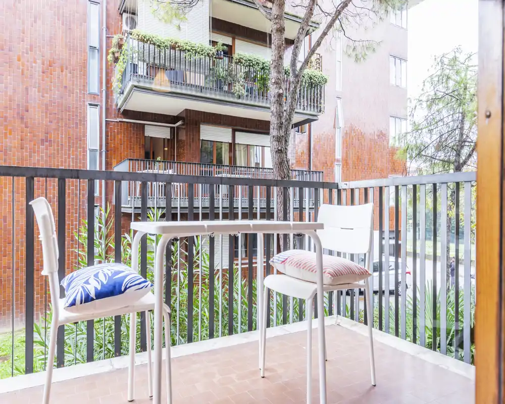 Bright furnished balcony with two chairs and a small table, overlooking a quiet courtyard and neighboring building — ideal for morning coffee.