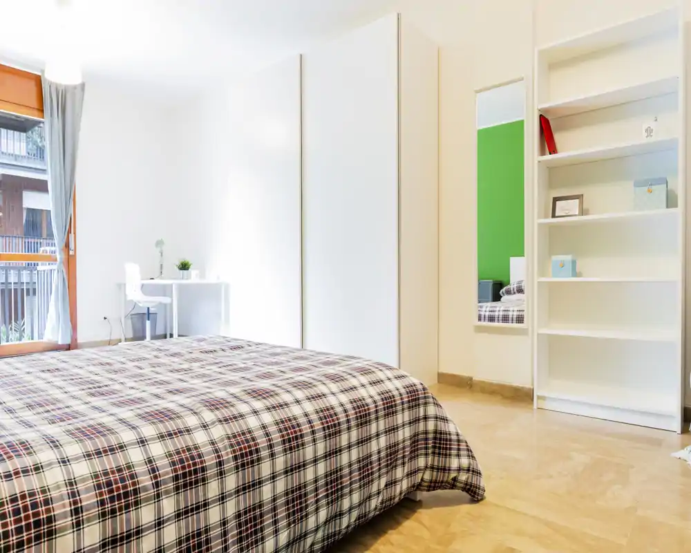 Bright, modern bedroom with a large bed covered in a checked duvet, a white wardrobe and a small desk by the window letting in natural light.
