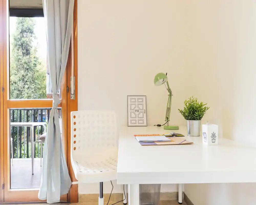 Bright, minimal living area with a white desk by a balcony door. The workspace features a modern white chair, green desk lamp and a small plant, ideal for remote work or study.